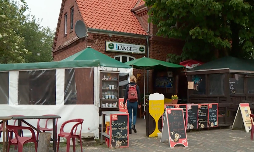 Eingang des Inselkaufmanns auf Neuwerk mit Backsteingebäude, grünen Sonnenschirmen, Kreidetafeln und kleinem Außenbereich.