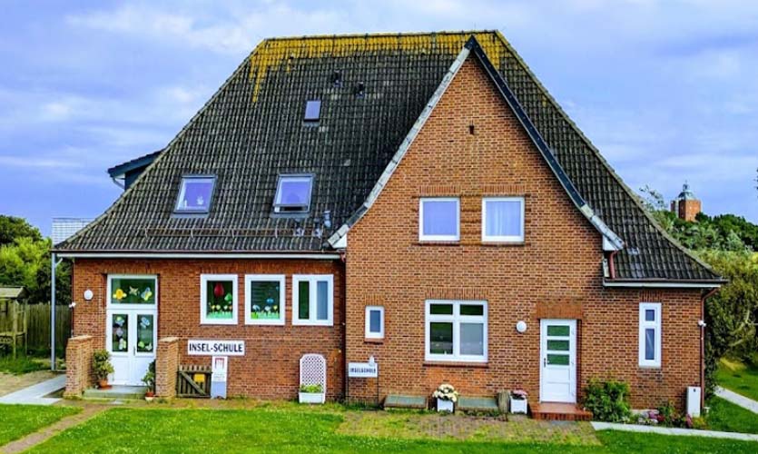 Backsteingebäude der Inselschule Neuwerk mit großem Dach, Eingangsschild, Fenstern und gepflegter Grünfläche davor.